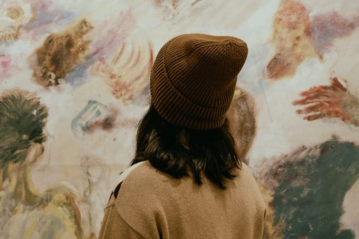 A person wearing a brown knit beanie and a beige jacket stands facing a colorful, abstract painting. The painting features various blended human-like forms and soft, mixed hues, creating a dreamlike effect. The person's hair is shoulder-length and dark.